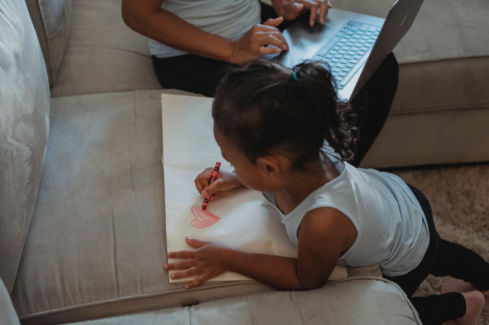 Child drawing a heart on paper while a parent works on a laptop at home, representing family visa holders working in the UAE